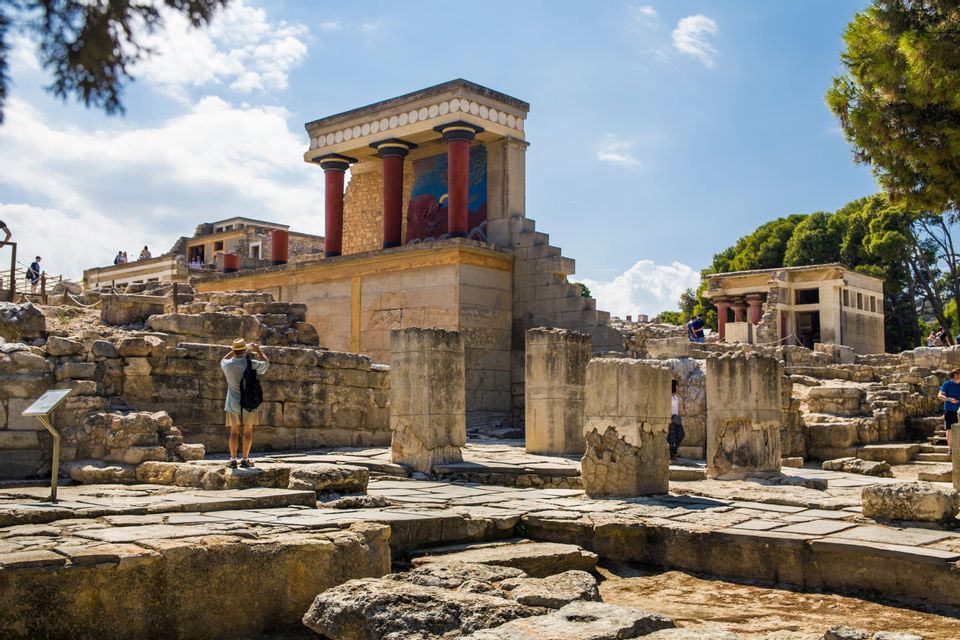 A person wearing a hat photographs the ancient stone ruins of a palace with red columns under a partly cloudy sky.