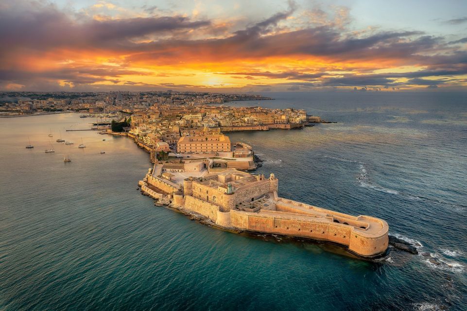 An aerial view of a historic coastal city and a stone fortress on a peninsula under a dramatic sunset sky.