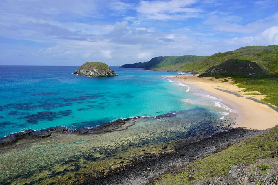 Una vista panoramica di una baia tropicale con acqua turchese, una spiaggia sabbiosa e colline verdi sotto un cielo azzurro con nuvole.