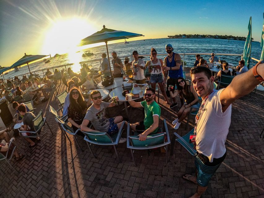 Un hombre se toma una selfie gran angular de su viaje en grupo de WeRoad en un bar concurrido frente al mar durante un atardecer vibrante.