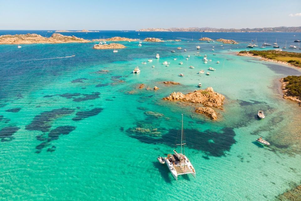 Vista aerea di un catamarano e altre barche ancorate in una baia turchese poco profonda tra isole rocciose.