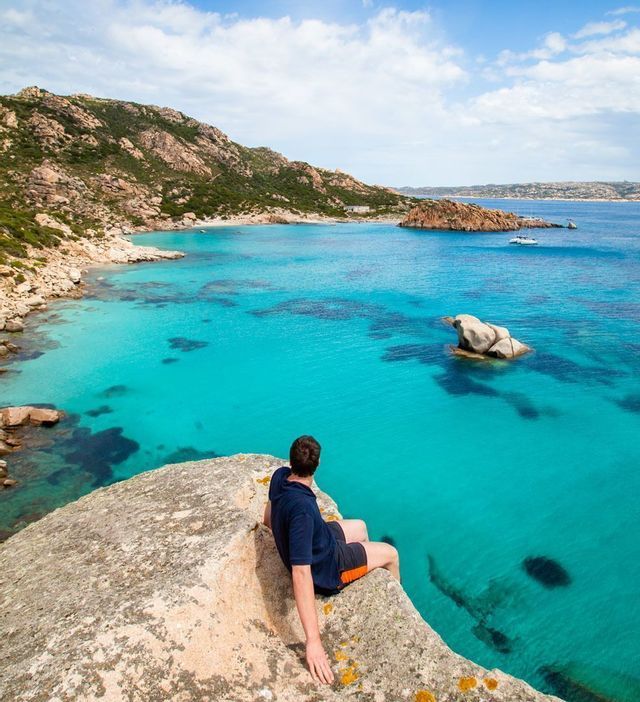 Un uomo in camicia blu siede su una grande roccia che domina una baia turchese e cristallina con una costa rocciosa.
