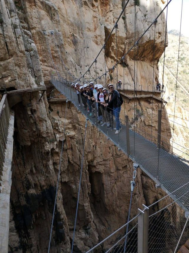 Eine WeRoad-Reisegruppe mit Helmen posiert für ein Foto auf einer Hängebrücke, die an einer steilen Canyonwand befestigt ist.