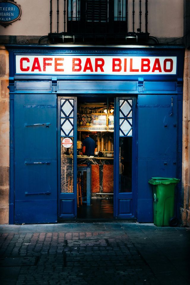 La vetrina blu del Cafe Bar Bilbao, con un'insegna illuminata e le porte aperte su una strada di ciottoli.