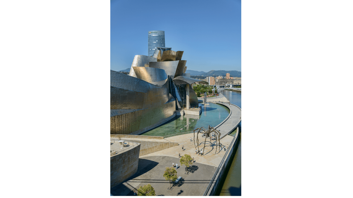 Una vista aerea di un moderno edificio metallico e una grande scultura di ragno accanto a un fiume sotto un cielo azzurro e limpido.