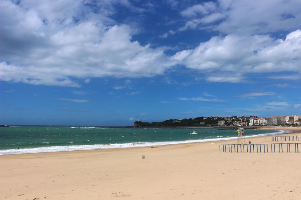Una spiaggia ampia e sabbiosa incontra il mare verde sotto un cielo blu con nuvole bianche, con una cittadina costiera in lontananza.