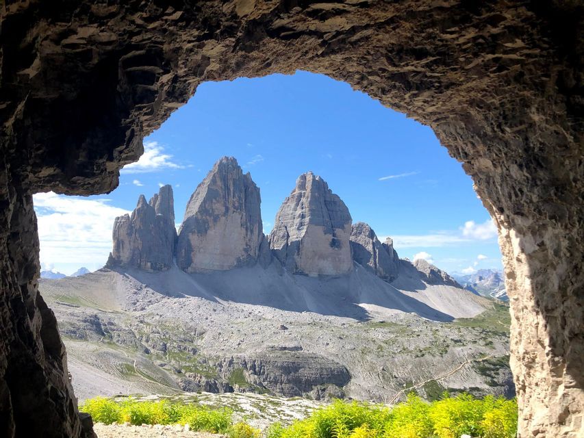 Pics montagneux déchiquetés vus depuis l'entrée d'une grotte rocheuse, avec des plantes jaunes vives au premier plan sous un ciel bleu clair.