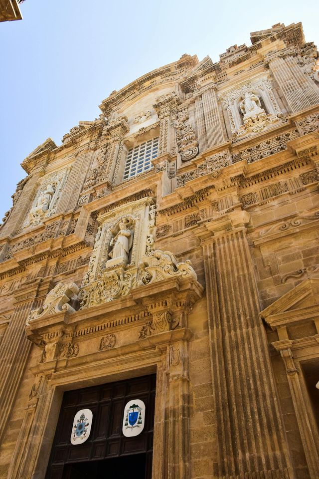 Una ripresa dal basso della facciata in pietra riccamente decorata di una chiesa barocca, con intagli complessi, colonne e statue, sotto un cielo limpido.