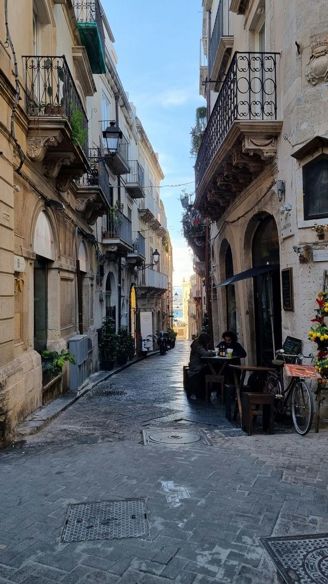 Vista su uno stretto vicolo acciottolato con persone in un caffè all'aperto, che porta al mare in lontananza.