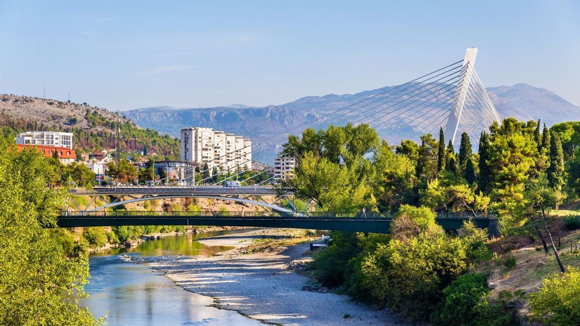 Un moderno ponte strallato bianco e un ponte pedonale verde attraversano un fiume poco profondo che scorre attraverso una città con montagne sullo sfondo.