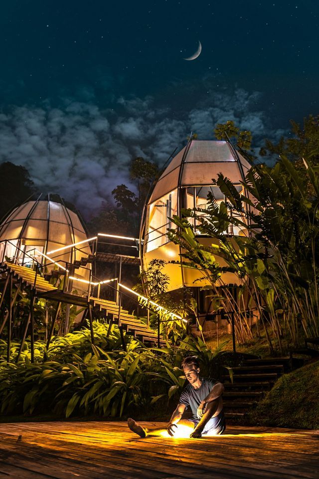 Un uomo siede su una terrazza di legno di notte, con una luce in mano davanti a due cabine a forma di cupola illuminate in una giungla lussureggiante.