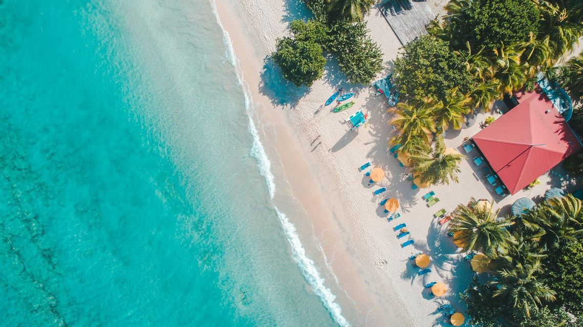 Vista aérea cenital de una playa tropical con agua turquesa, arena blanca, palmeras, kayaks de colores y un edificio con tejado rojo.