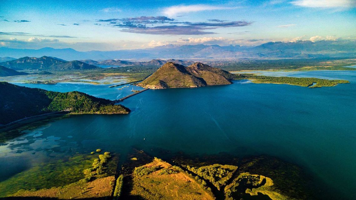 Una veduta dall'alto di un immenso lago blu, circondato da verdi terre, con un ponte che collega una penisola e le montagne in lontananza.