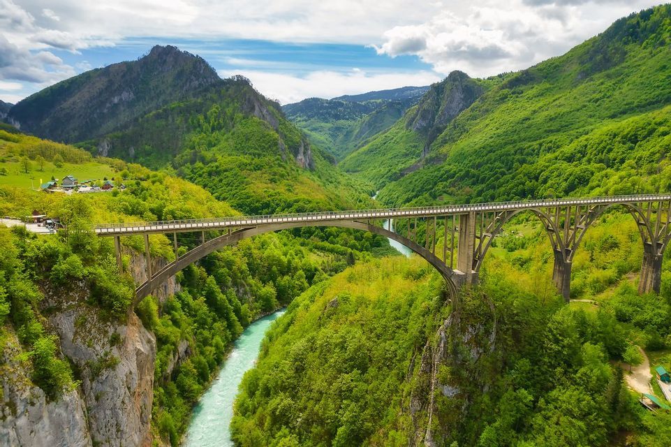 Un ponte ad arco attraversa un fiume turchese in una profonda valle, circondata da lussureggianti montagne verdi, sotto un cielo parzialmente nuvoloso.