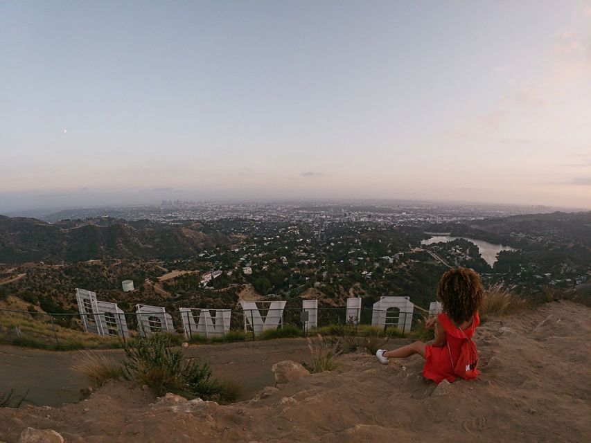 Une femme en robe rouge est assise sur une colline derrière le panneau Hollywood, dominant un vaste paysage urbain au crépuscule.