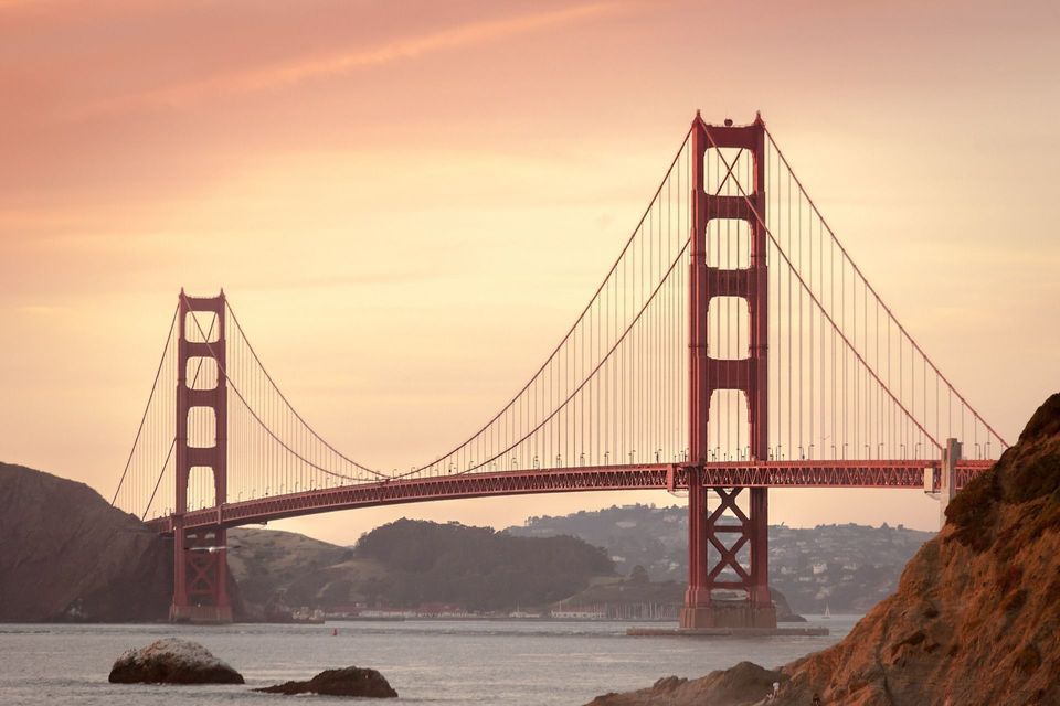 Eine große rote Hängebrücke überspannt ein Gewässer mit Hügeln im Hintergrund unter einem warm getönten Himmel bei Sonnenuntergang.