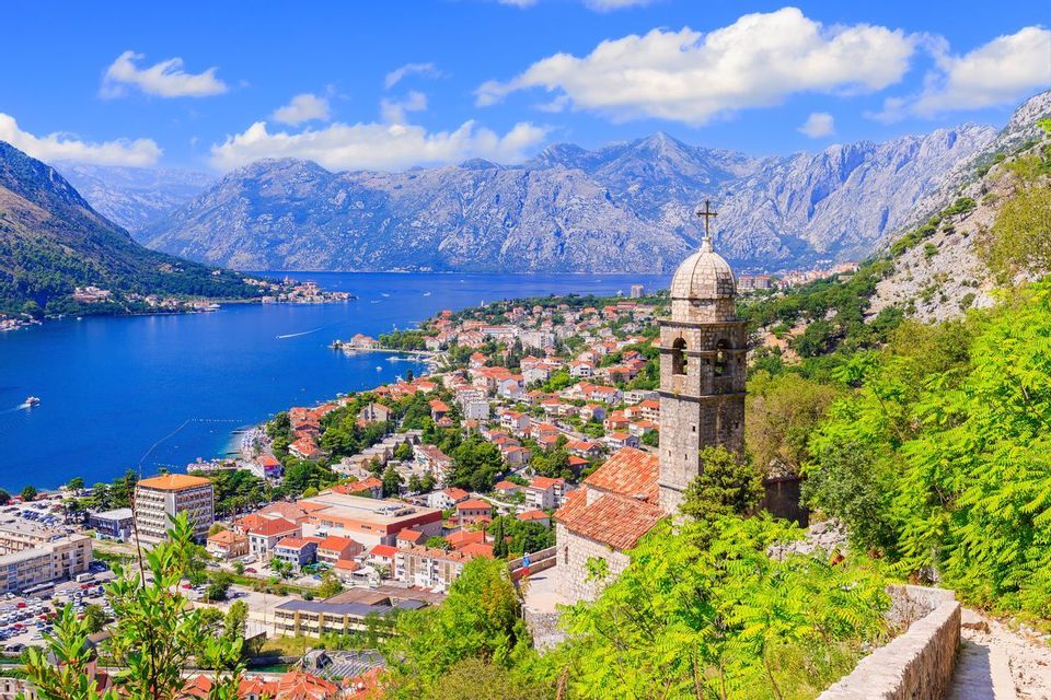 Un campanile in pietra domina una città costiera incastonata tra una baia azzurra e imponenti montagne sotto un cielo soleggiato.