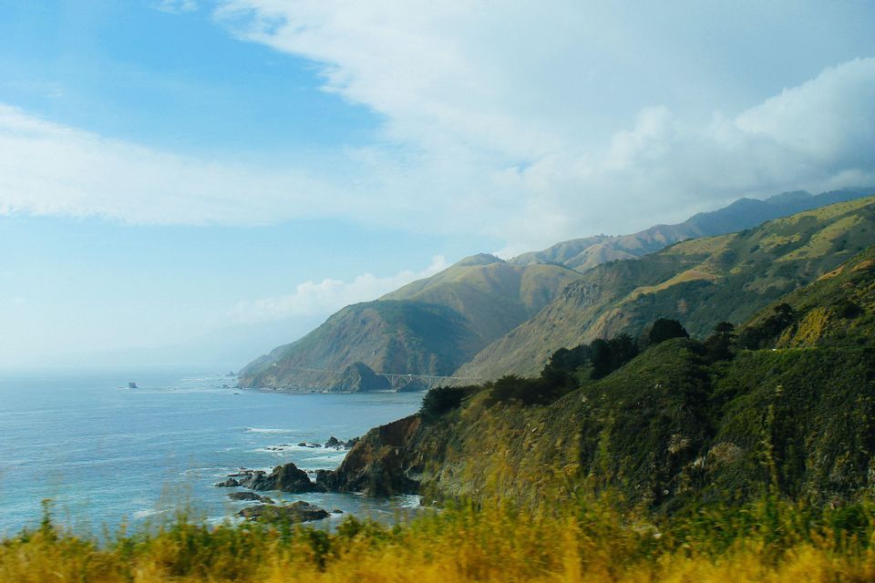 Una vista de una costa escarpada con montañas verdes que descienden hacia el océano azul, bajo un cielo parcialmente nublado y con un puente a lo lejos.