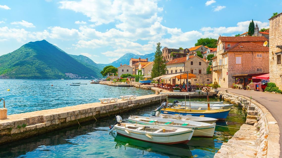 Piccole imbarcazioni sono ormeggiate lungo un molo di pietra in una cittadina di mare, con verdi montagne che si ergono sullo sfondo sotto un cielo blu, parzialmente nuvoloso.