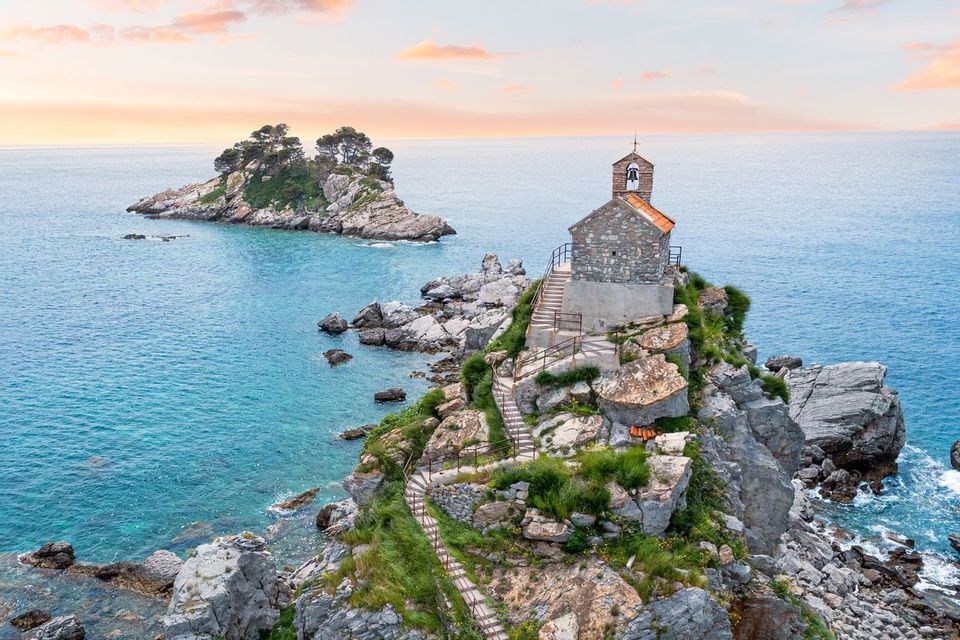Una vecchia chiesa in pietra con un campanile si erge su un isolotto roccioso nel mare, vista al tramonto.