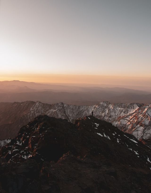 Una persona en silueta se alza sobre la cima de una montaña rocosa, contemplando una vasta cordillera nevada bajo un suave cielo de amanecer naranja.