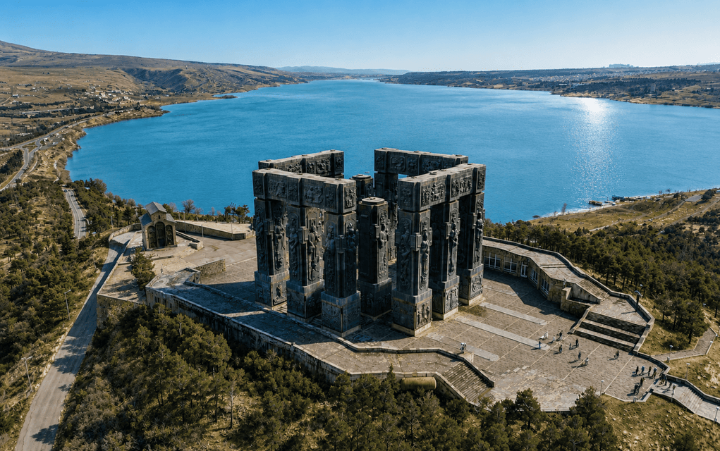 Veduta aerea di un grande monumento in pietra scura, situato su una piazza collinare, con vista su un vasto lago blu sotto un cielo limpido.