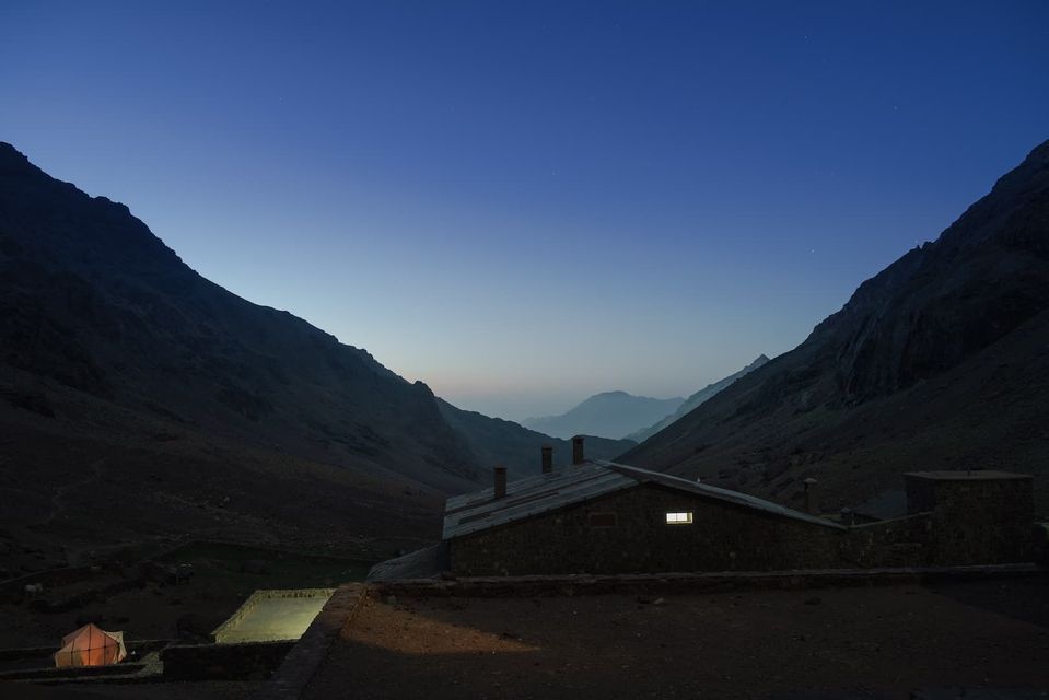 Un edificio de piedra con una ventana iluminada y una tienda de campaña iluminada enclavada en un valle de montaña al anochecer bajo un cielo azul claro.