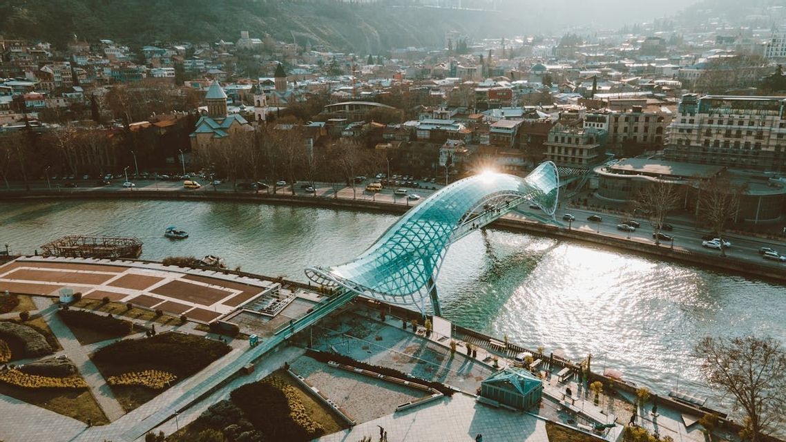 Una veduta aerea di un moderno ponte pedonale in vetro e acciaio che si estende su un fiume, con uno skyline cittadino sullo sfondo.