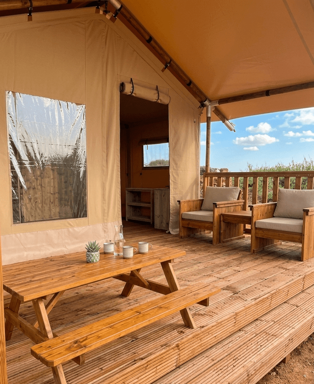 La pedana in legno di una tenda glamping in tela, arredata con un tavolo da picnic e due poltrone sotto una tenda da sole.