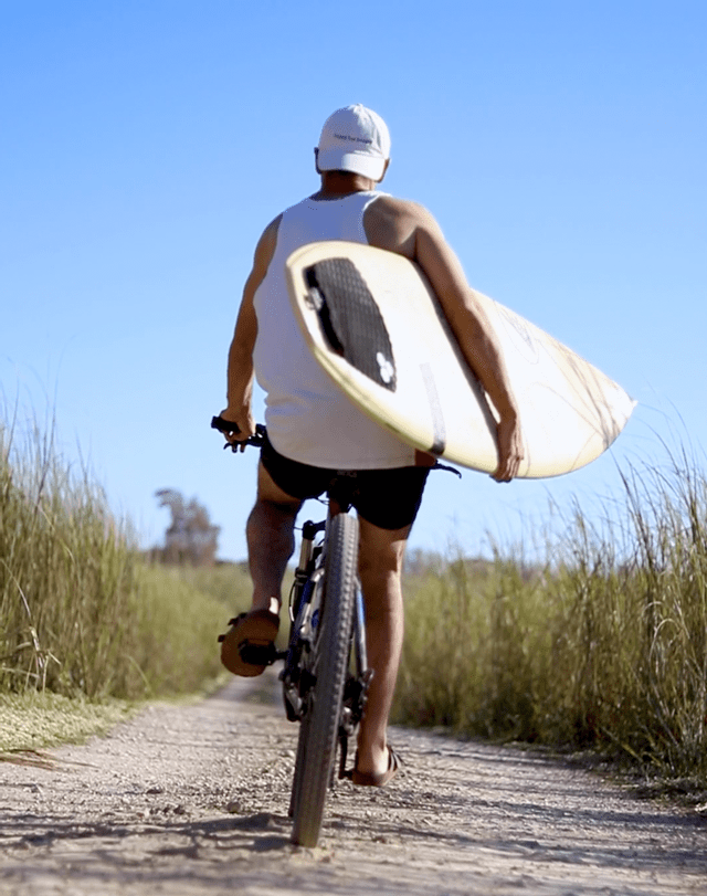 Un uomo visto di spalle pedala in bicicletta su un sentiero sterrato, con una tavola da surf sotto il braccio.