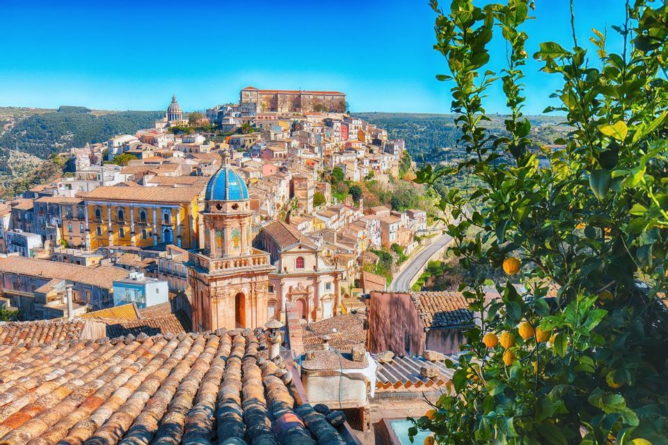 Vista soleggiata dall'alto di una città storica su una collina con una cupola blu, tra tetti in terracotta e un albero di limoni.