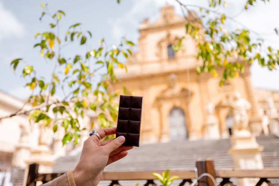 Una mano regge una tavoletta di cioccolato fondente in primo piano, con un edificio ornato e color sabbia sfocato sullo sfondo.