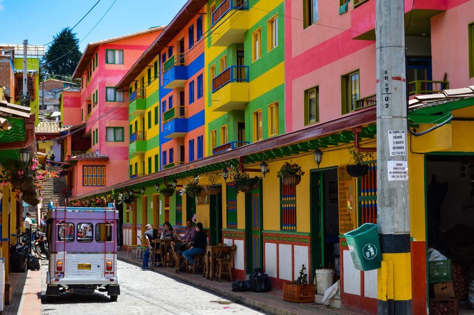 A cobblestone street lined with brightly colored, striped buildings, where a purple vehicle is parked and people sit at outdoor tables.