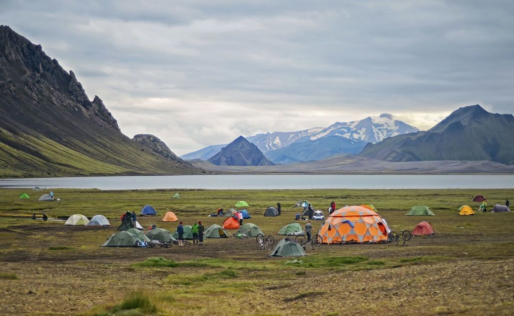 Ein großer Campingplatz mit farbenfrohen Zelten und Fahrrädern auf einer grasbewachsenen Ebene neben einem See, mit schroffen Bergen im Hintergrund unter einem bewölkten Himmel.