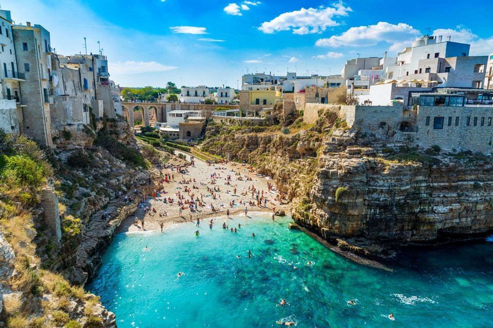 Una vista dall'alto di una spiaggia affollata in una caletta con acqua turchese, circondata da scogliere sormontate da edifici.
