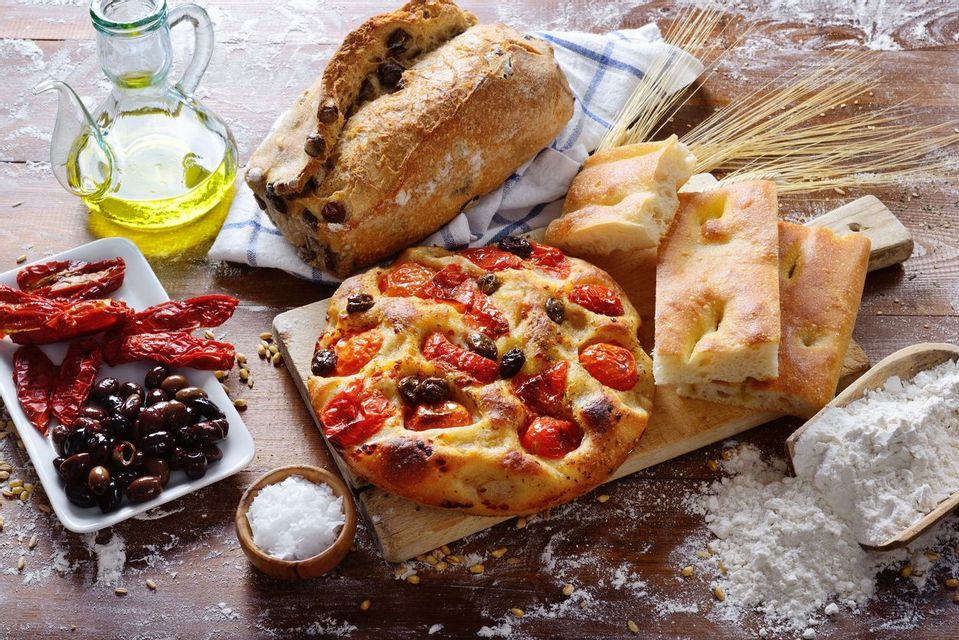 Un'ampia selezione di pani italiani, inclusi focaccia e pane alle olive, con ingredienti come olio d'oliva, pomodori e farina, su un tavolo di legno.