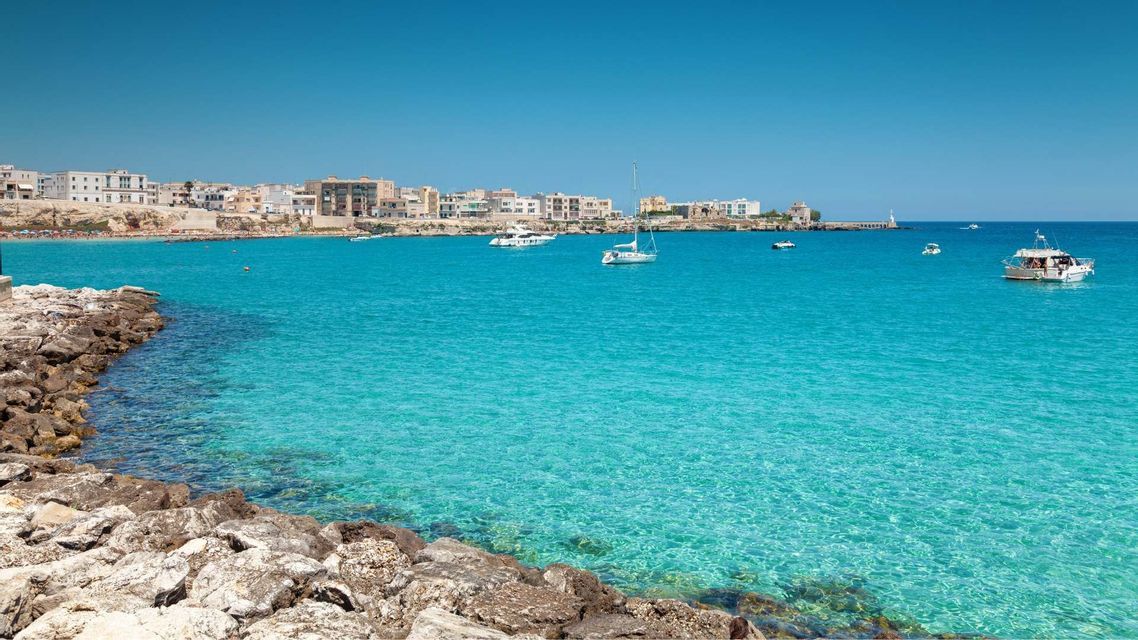 Una costa rocciosa si affaccia su una baia turchese con diverse barche ancorate vicino a una località costiera.