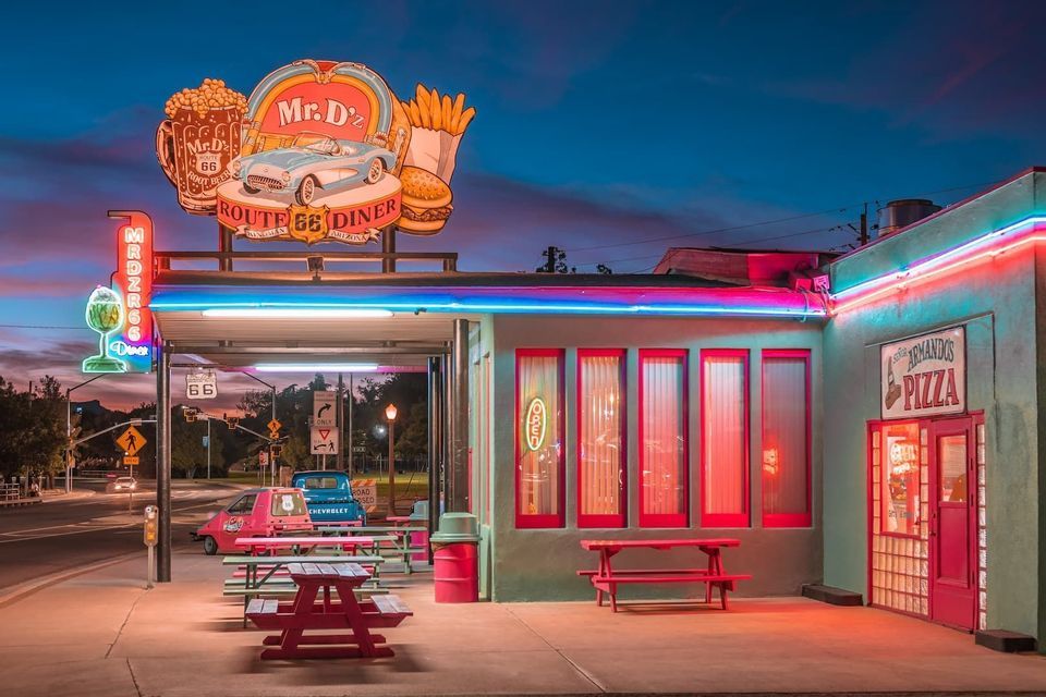 Un diner americano in stile retrò illuminato da insegne al neon colorate al tramonto, con tavoli da picnic all'aperto.