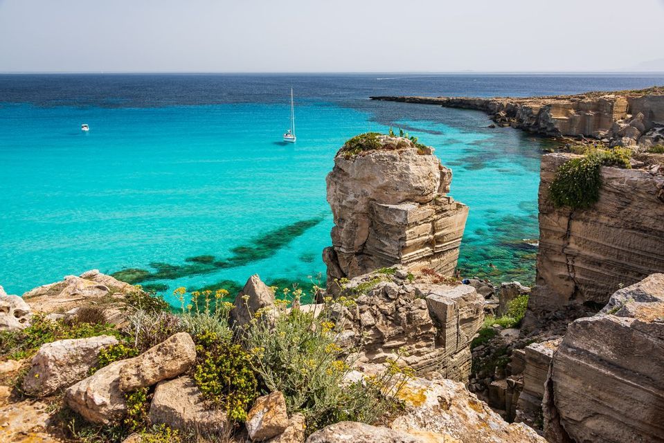 Vista da una scogliera rocciosa su una baia turchese, con una barca a vela vicino a un'imponente formazione rocciosa.