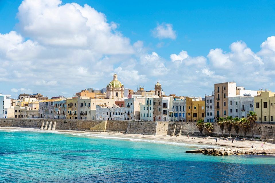 Una città costiera con edifici colorati si erge dietro un muro di pietra, affacciata su un mare turchese e una spiaggia sabbiosa sotto un cielo azzurro.