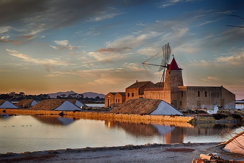 Un mulino a vento tradizionale con tetto rosso si erge accanto alle saline sul bordo dell'acqua al tramonto, con montagne in lontananza.