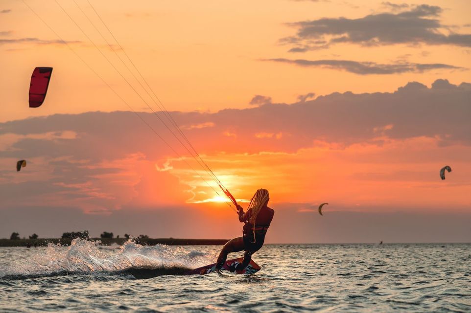 Una persona con lunghe trecce fa kitesurf sul mare, stagliata contro un vibrante tramonto arancione.