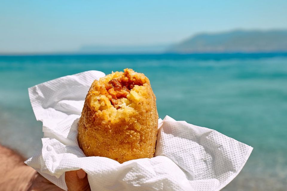 Una mano tiene un arancino di riso fritto ripieno di carne in un tovagliolo, con il mare blu sullo sfondo.