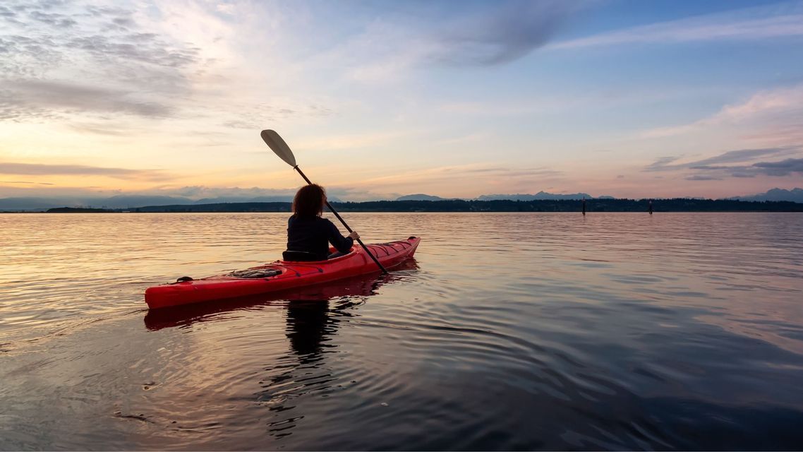 Una persona vista di spalle pagaia su un kayak rosso su acque tranquille, osservando il tramonto sulle montagne distanti.