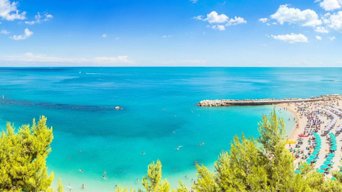 Una veduta aerea di una spiaggia affollata con file di ombrelloni lungo il mare calmo e turchese in una giornata di sole.