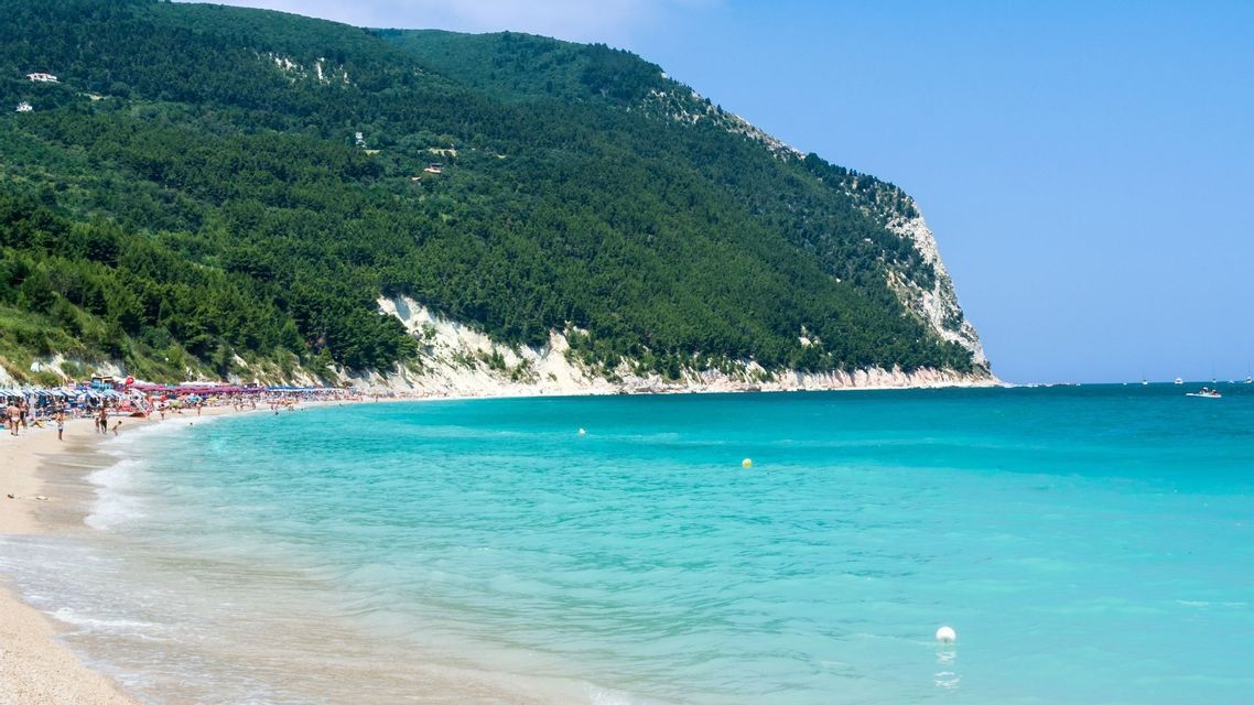 Una spiaggia sabbiosa affollata ai piedi di una montagna alberata, con acqua turchese che lambisce la riva sotto un cielo azzurro e limpido.