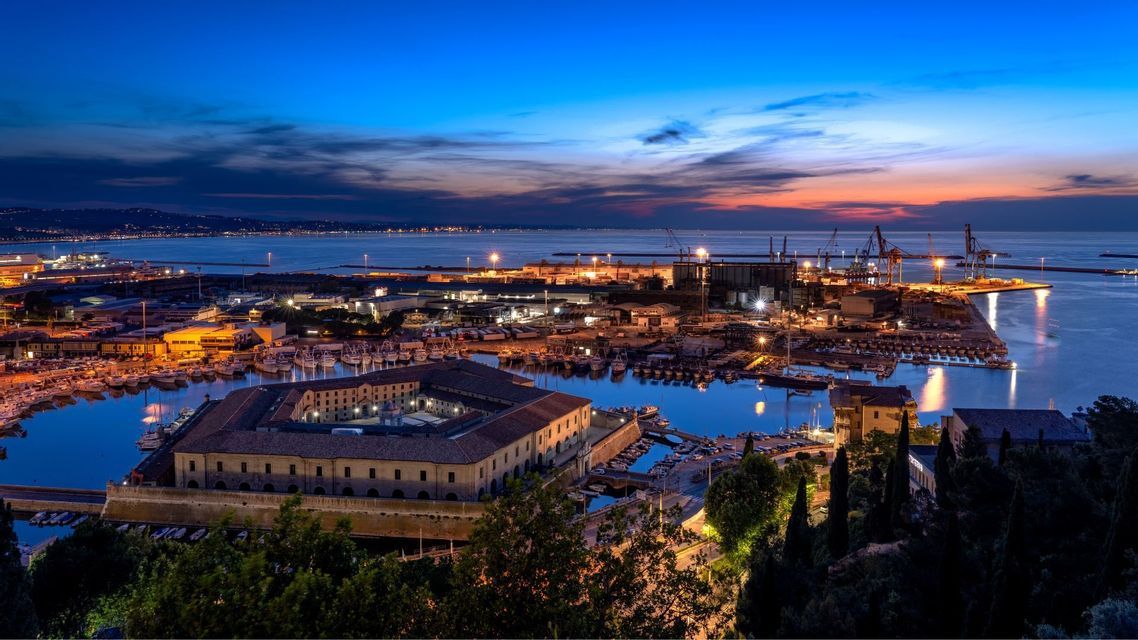 Una vista dall'alto di una grande città portuale illuminata al tramonto, con una fortezza storica nel porto.