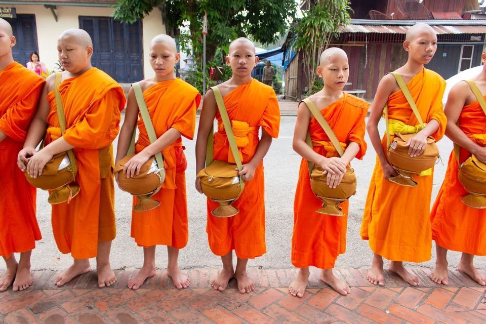 Eine Reihe junger buddhistischer Mönche in orangefarbenen Roben steht barfuß auf einer Ziegelstraße, jeder mit einer Almosenschale.