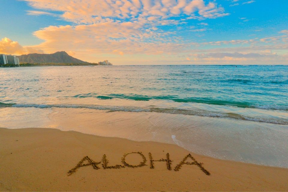 ALOHA im Sand am Strand, mit Wellen und Bergblick bei Sonnenuntergang.