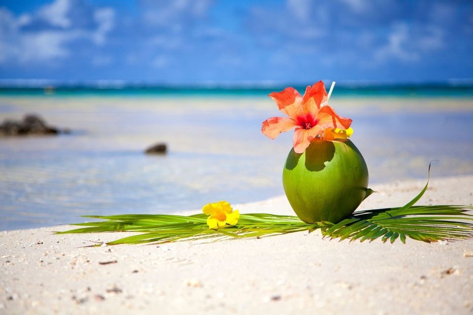 Ein Kokosnuss-Cocktail, garniert mit einer orangen Hibiskusblüte, steht an einem weißen Sandstrand mit dem Ozean im Hintergrund.